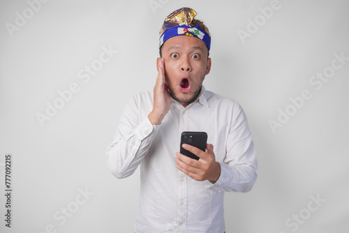 Young Balinese man wearing white shirt and traditional headdress showing shocked expression on face while holding smartphone, surprised after reading news or gossip, isolated by white background © Reezky