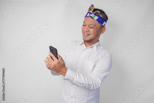 Excited Balinese man wearing white shirt and traditional headdress smiling happily while using smartphone, replying message, reading news © Reezky