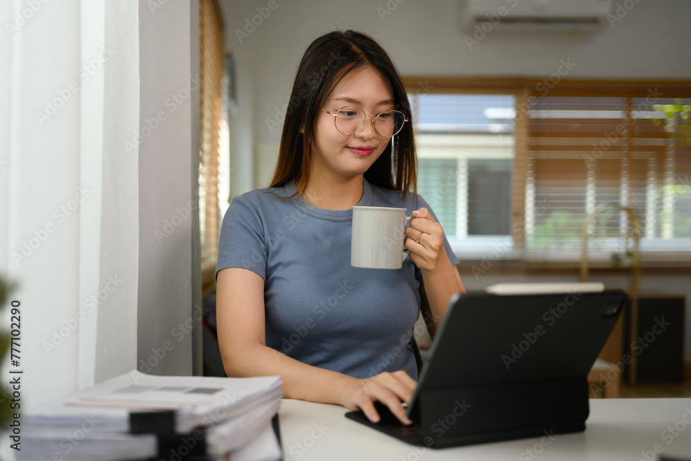 Smiling Asian woman drinking coffee and working on digital tablet at desk in living room Smiling Asian woman drinking coffee and working on digital tablet at desk in living room