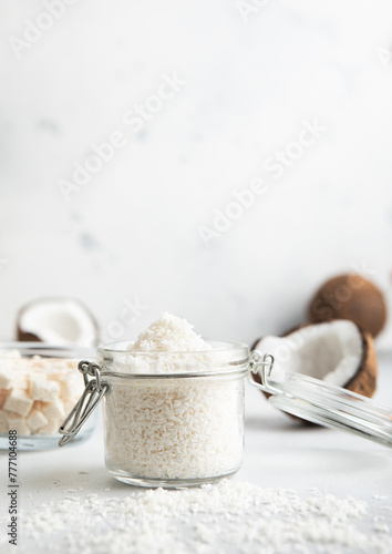 Shredded sweet coconut flakes in glass jar with ripe coconuts on light table.
