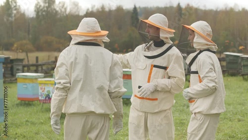 Medium shot of three professional beekeepers wearing protective clothing having conversation at apiary, discussing plan of work