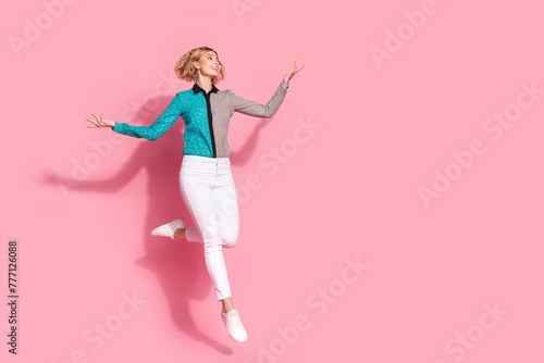 Full size photo of pretty young girl jumping raise hands look empty space wear trendy striped cyan outfit isolated on pink color background