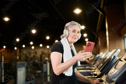 jolly mature woman with towel on shoulders and headphones holding phone and exercising on treadmill photo