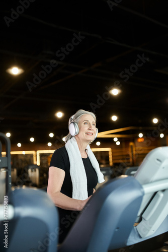 joyful pretty senior woman in cozy attire training on treadmill and listening music in headphones