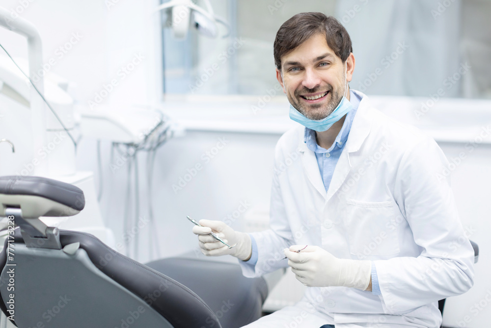 Professional male stomatologist at workplace in clinic interior, dentist doctor in uniform with tools in hands smiling at camera, ready for check up Professional male stomatologist at workplace in clinic interior, dentist doctor in uniform with tools in hands smiling at camera, ready for check up