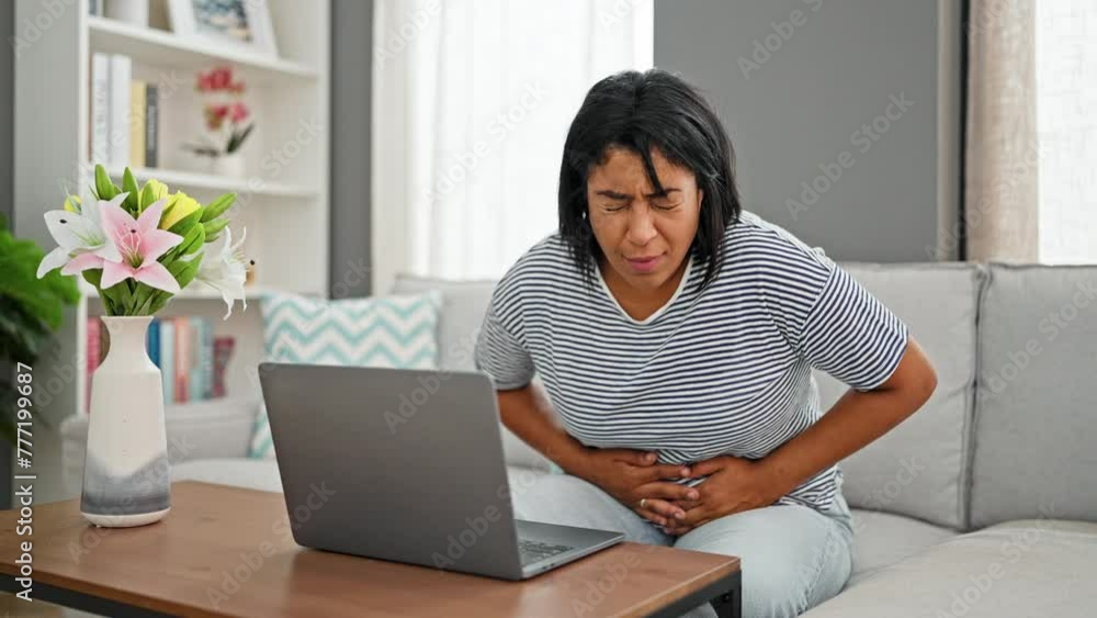 Hispanic woman working on laptop at home, feeling abdominal pain, in a modern living room. Hispanic woman working on laptop at home, feeling abdominal pain, in a modern living room.