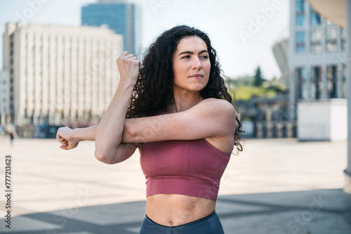 Waist portrait of athletic young woman warming up, stretching arms outdoors