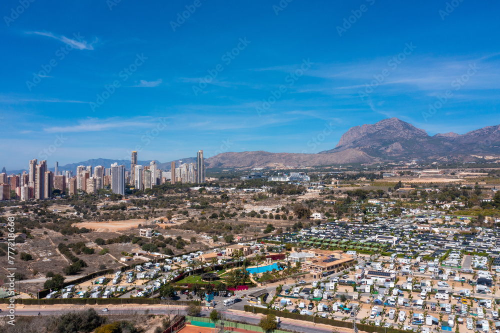 Aerial photo of the town of Benidorm in Spain showing a drone view of a camp site with many motorhomes and caravans on the camping site on a sunny day with mountains and apartments in the background