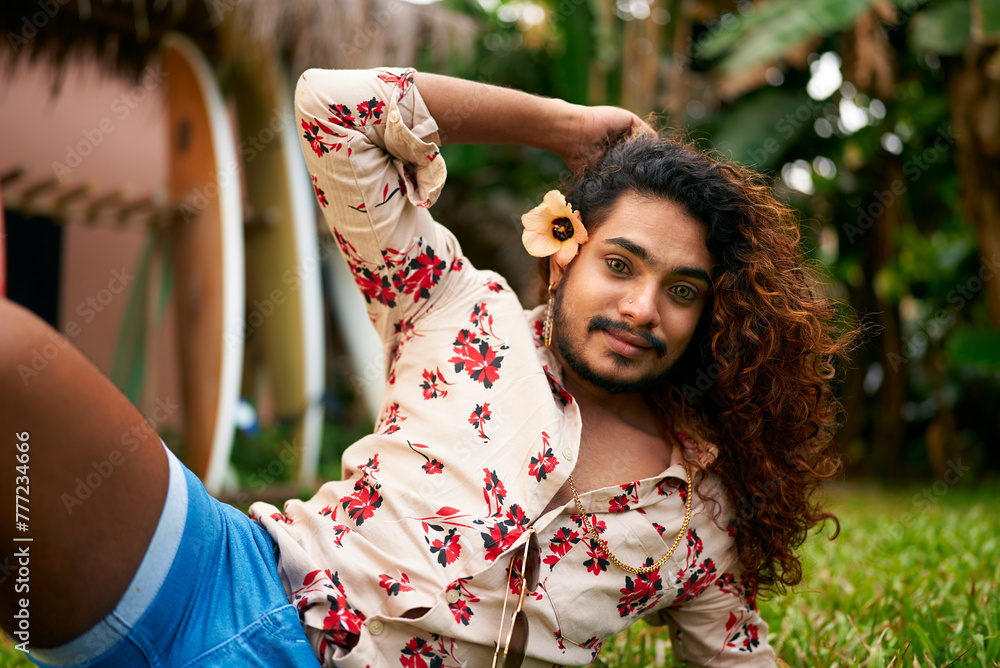 Obraz premium Gay man with floral shirt poses, holds flower, exudes confidence, style in rich garden. Vibrant outdoor setting for travel, lifestyle blogs. LGBT-friendly tropical vacation scene.