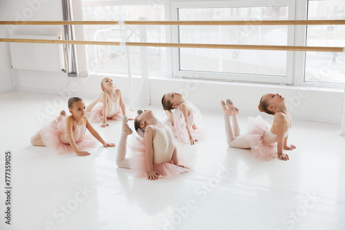 Group of little girls ballerinas doing floor stretches in a bright modern dance studio. Classical ballet school. Concept of art, sport, education, hobby, active lifestyle, leisure time. photo