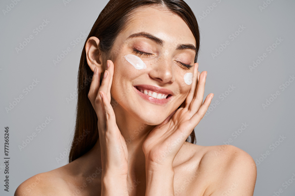 A young Caucasian woman with brunette hair smiles brightly while delicately touching her face with cream