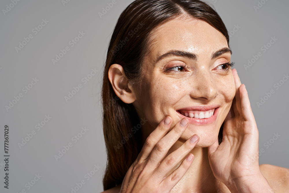A young Caucasian woman with brunette hair smiles warmly, gently touching her face with her hands in a studio setting. A young Caucasian woman with brunette hair smiles warmly, gently touching her face with her hands in a studio setting.
