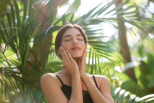 A beautiful woman with her eyes closed stands in front of green plants and palm trees, holding both hands to her face for skin care