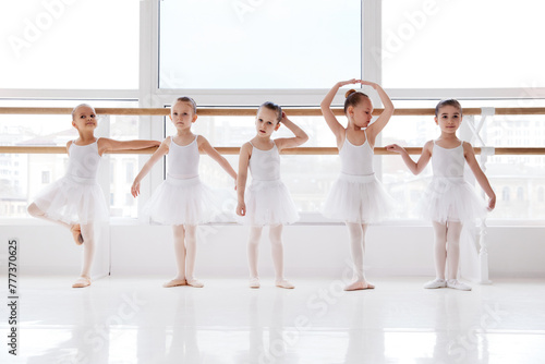 Group of children, school-girls in white tutu training poses near barre in light room. Classical ballet school. Concept of art, sport, education, hobby, active lifestyle, leisure time.