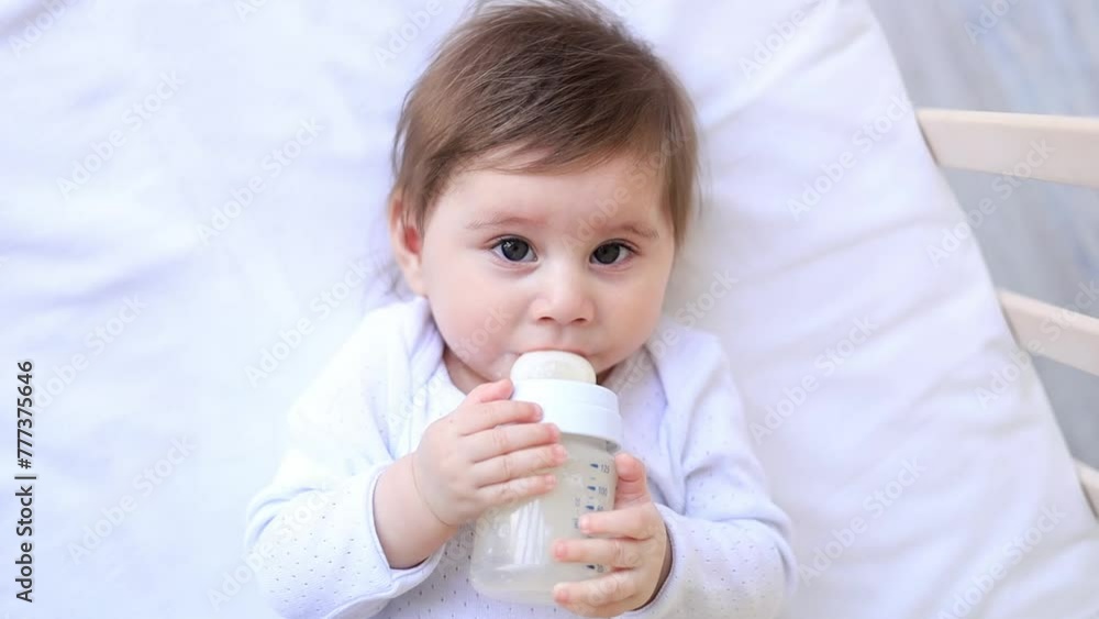 Baby newborn girl sucks a bottle of milk lying on her back in a crib in white clothes, baby food concept, close-up Baby newborn girl sucks a bottle of milk lying on her back in a crib in white clothes, baby food concept, close-up