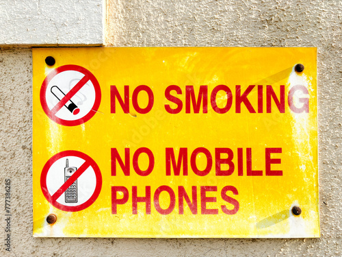 Front view of a yellow sign board displayed on a cream color wall saying 'on smoking' and 'no mobile phones' with it's symbols on the left side.