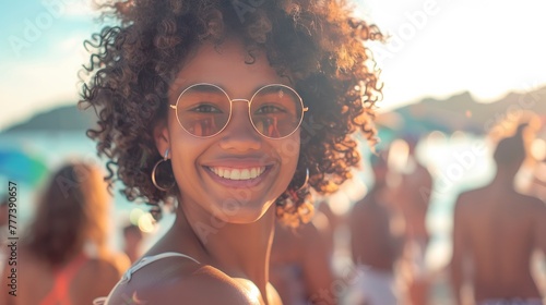 A beautiful mixed race woman with curly hair smiling at the camera in sunglasses on the beach, surrounded by people having fun during a Summer festival, sunlight and a bokeh effect,