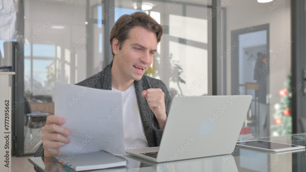 Young Man Celebrating while Using Laptop and Documents Young Man Celebrating while Using Laptop and Documents