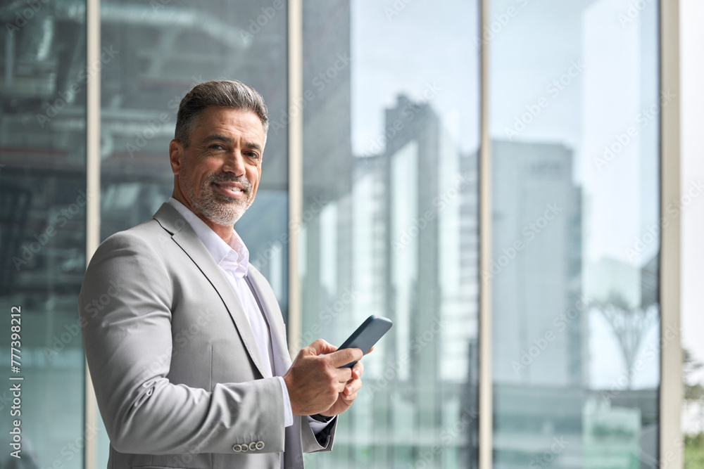 Happy rich middle aged businessman using cellphone, busy older business man leader investor, mature male executive in suit holding mobile phone standing at office window looking at camera, portrait. Happy rich middle aged businessman using cellphone, busy older business man leader investor, mature male executive in suit holding mobile phone standing at office window looking at camera, portrait.