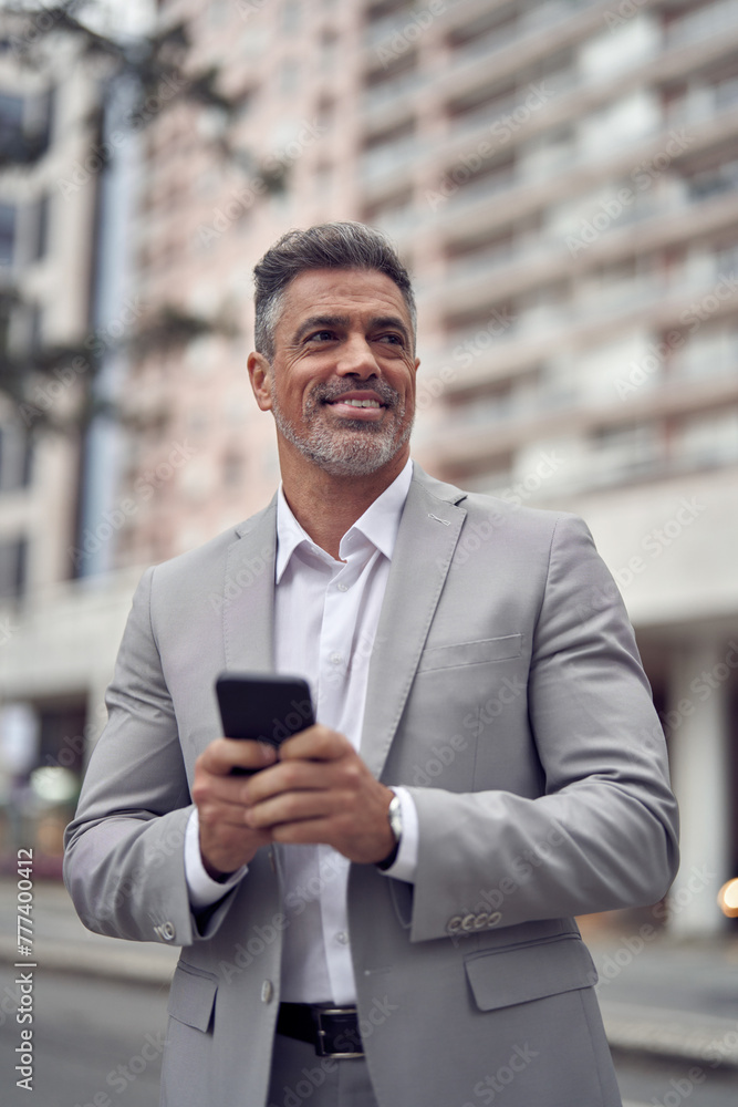 Happy middle aged business man entrepreneur investor standing outside office building using mobile phone looking away. Mature businessman professional executive holding smartphone, vertical. Happy middle aged business man entrepreneur investor standing outside office building using mobile phone looking away. Mature businessman professional executive holding smartphone, vertical.