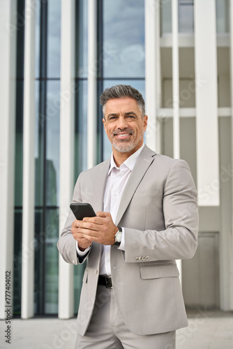 Confident happy middle aged banker, smiling mature business man corporate leader investor entrepreneur wearing suit standing outdoors using smartphone with mobile phone in hands. Vertical portrait.