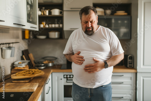 A man who is overweight is standing in the kitchen and has stomach pains or abdominal pain and rubs his stomach in pain with his hands