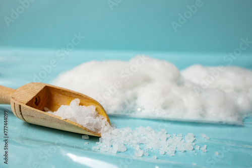 Rock salt in wooden scoop next to white foam blob on blue background, soft focus close up