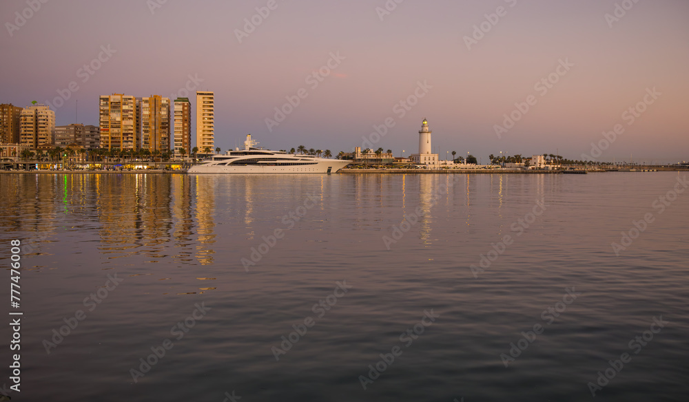 Panoramic view with the first lights and its reflections in the water, of lighthouse called La Farola, pier 1 and a yacht. Malaga, Spain