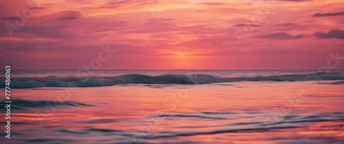 A pink sunset over the ocean, with waves gently lapping at shore and a distant sun setting behind dark clouds. The sky is painted in shades of orange and purple as it sets on horizon. 