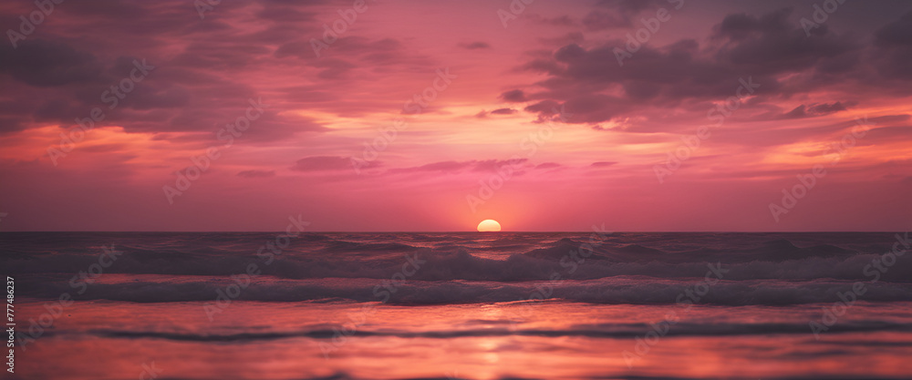 A pink sunset over the ocean, with waves gently lapping at shore and a distant sun setting behind dark clouds. The sky is painted in shades of orange and purple as it sets on horizon. 