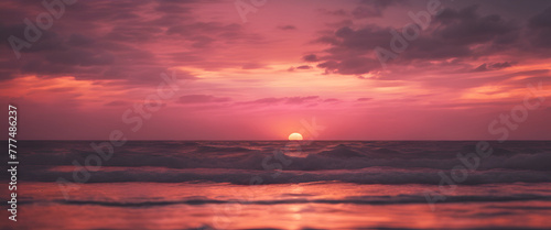 A pink sunset over the ocean  with waves gently lapping at shore and a distant sun setting behind dark clouds. The sky is painted in shades of orange and purple as it sets on horizon. 