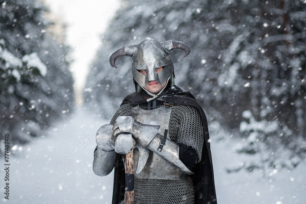 Fototapeta premium Portrait of a medieval fantasy warrior in a horned helmet, steel cuirass, chain mail with a two-handed ax in his hands, posing against the backdrop of a winter forest. 