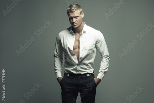 Male fashion, beauty concept. Portrait of brutal young man with short wet blond hair wearing white shirt, black pants, posing over gray background. Classic style. Text space. Studio shot