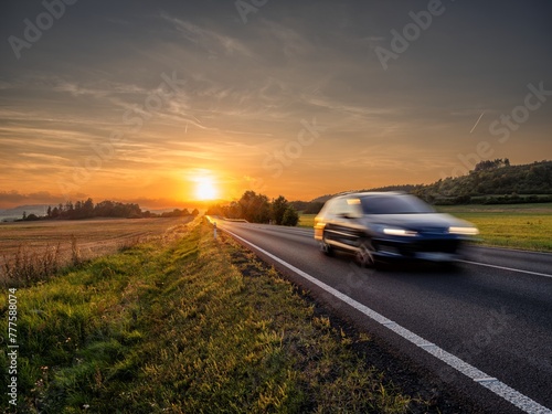 Motion blurred car driving on the asphalt road in rural landscape at sunset