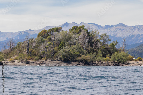 Horizontal photo of mountains full of vegetation and rocks, surrounded by tranquil waves of a lake's water, with a clear blue sky during a sunny day, in Lago Nahuel Huapi, Argentina