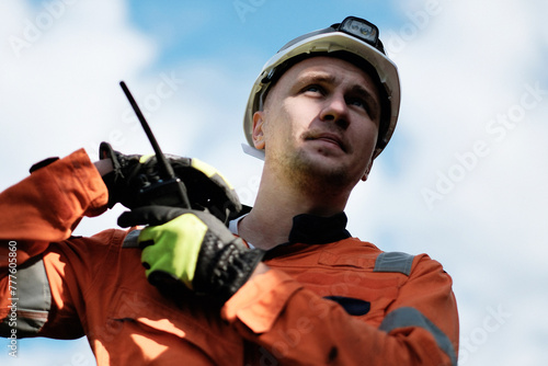 Wind Farm Offshore Maintenance Technician. Seafarer. Seaman. Navigator. A Man In A Working Overall Boiler Suit With A Radio And Safety Helmet With A Blurred Wing Generators In The Background. photo