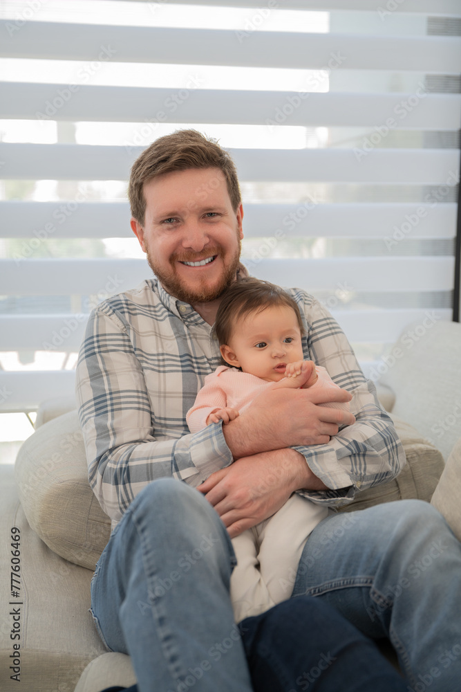 Fototapeta premium Portrait of dad holding his baby daughter in his arms, It's daytime he looks at the camera sitting on the sofa.