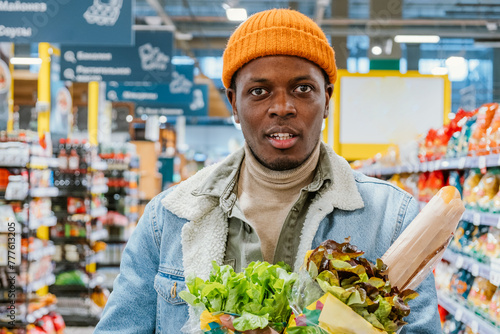 Young African American Man Shopping for Fresh Produce in Grocery Store. A man in a trendy orange beanie and denim jacket holds fresh bread and leafy greens while shopping in a grocery store