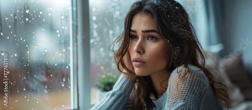 A woman with a melancholic expression observes rain falling outside the window.