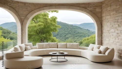 A stylish seating area with a curved cream-colored sofa adorned with plush pillows, set against a backdrop of smooth stone walls and oversized windows offering panoramic views of lush greenery outside