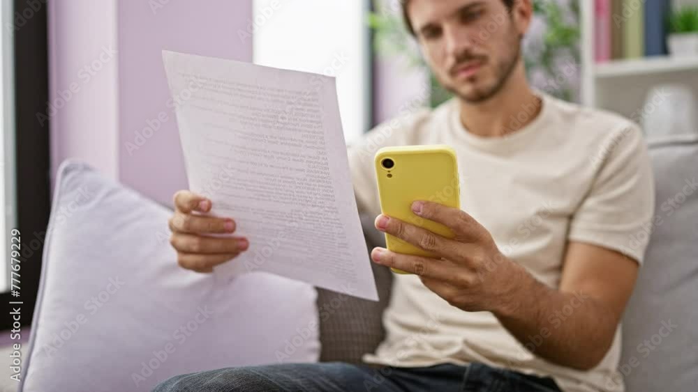 A focused young man reads a document while holding a smartphone at home, showcasing multitasking in a domestic setting. A focused young man reads a document while holding a smartphone at home, showcasing multitasking in a domestic setting.