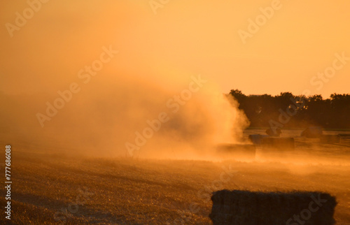 Combine harvester pressing straw from field into bales driving field on sunny summer evening. Field with bales of pressed wheat. Lots of dust on field. Agricultural agro industrial harvesting works