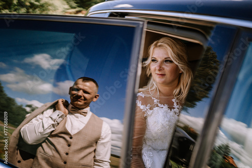 Valmiera, Latvia - Augist 13, 2023 - A bride inside a car is captured in focus, while the groom's reflection is seen on the car's surface as he adjusts his vest. photo