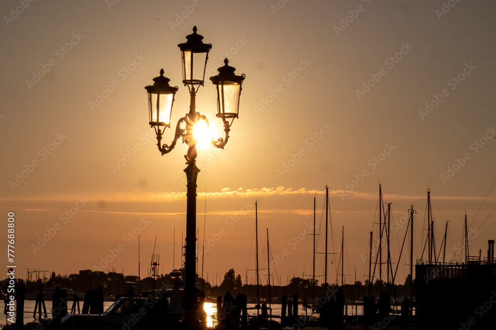 Silhouette of street light and  sailing boats in port of  charming town of Chioggia at sunrise, nestled in the stunning Venetian Lagoon of the Veneto region in Italy. Tranquil atmosphere in summer