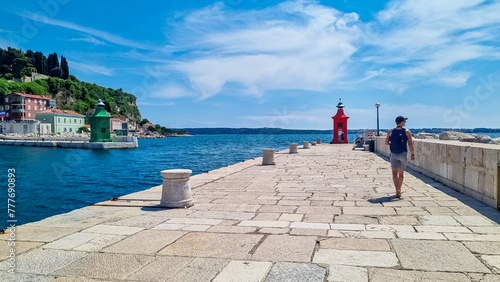 Tourist man walking along picturesque harbor of coastal town Piran. Walking next to the lighthouses overlooking the Adriatic Mediterranean Sea. Summer scene in small port.