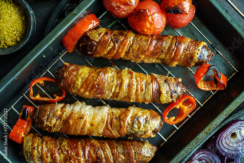 Grilled bacon wrapped sausages with tomatoes and onions on baking tray top view, close-up