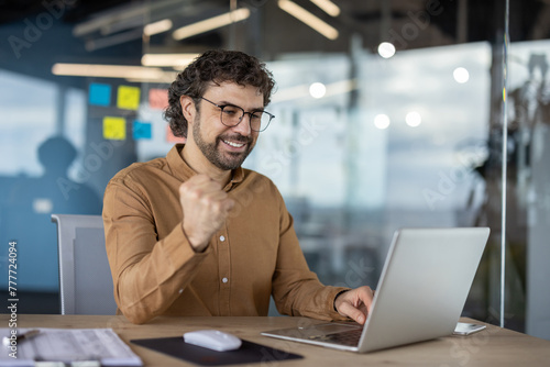 Cheerful businessman with glasses celebrating a success while working on his laptop in a modern office environment.