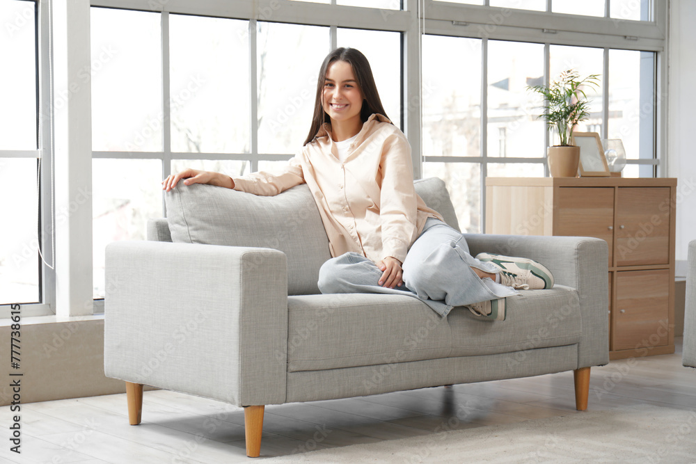 Pretty young woman sitting on grey sofa in living room - obrazy, fototapety, plakaty 