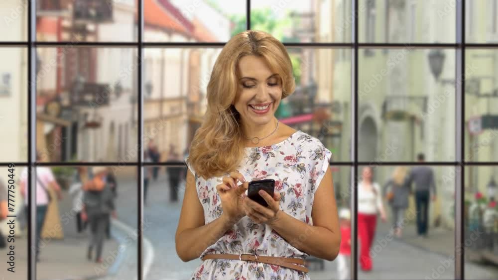 Portrait of a blonde woman using her smartphone indoors. Window with city street view in the background. Portrait of a blonde woman using her smartphone indoors. Window with city street view in the background.