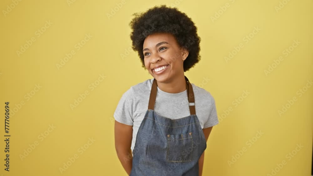 Laughing, confident african american woman with natural expression, wearing apron, standing and looking away to side with a smile on yellow isolated background